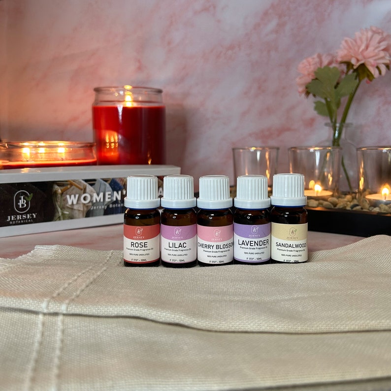 May include: Five small glass bottles with white lids and labels. The bottles are filled with essential oils and labeled with the scents: Rose, Lilac, Cherry Blossom, Lavender, and Sandalwood. The bottles are arranged in a row on a white surface. The text "WOMEN'S" is visible in the background.