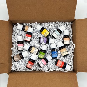 May include: A cardboard box filled with small glass bottles of essential oils. The bottles are labeled with different scents, including Rose, Cherry Blossom, Cappuccino, Sandalwood, Lemon, Grape, Cherry, Fresh Linen, Fabric Softener, Apple, Blueberry, Cinnamon, Thousand Wish, and Strawberry.