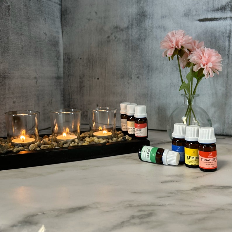 May include: A set of six essential oil bottles with labels for rose, lemongrass, lemon, strawberry, and grass. The bottles are arranged on a wooden tray with three lit tea lights.