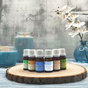 May include: Seven small glass bottles with white lids and colorful labels containing essential oils. The labels read "Grass", "Fabric Softener", "Blueberry", "Fresh Linen", "Nickerdoo", and "Apple". The bottles are arranged on a wooden slice with two blue candles in the background.