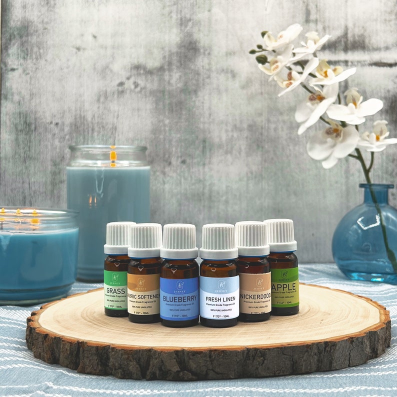 May include: Seven small glass bottles with white lids and colorful labels containing essential oils. The labels read "Grass", "Fabric Softener", "Blueberry", "Fresh Linen", "Nickerdoo", and "Apple". The bottles are arranged on a wooden slice with two blue candles in the background.