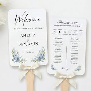 Könnte beinhalten: Zwei weiße Hochzeitsfächer mit Holzgriffen und cremefarbenen Schleifen. Die Fächer zeigen Blumendesigns und den Text "Welcome" und "The Ceremony". Das Hochzeitsdatum ist der 21.08.2026. Die Fächer sind für eine Hochzeit.