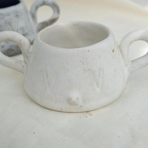 Può includere: Una piccola tazza in ceramica bianco sporco con due manici arrotondati. La tazza ha una consistenza macchiettata e una piccola sporgenza arrotondata sul davanti. La tazza è appoggiata su una superficie bianca.