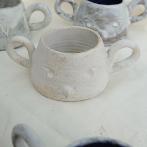 Può includere: Una tazza in ceramica fatta a mano con un design a forma di viso stravagante. La tazza bianco sporco presenta due manici e un piccolo viso rotondo con dettagli di occhi e naso. Altre tazze simili sono visibili sullo sfondo.