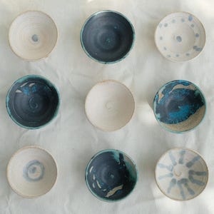 May include: A collection of nine handcrafted ceramic bowls. The bowls display a range of colours, including white, blue, and teal, with diverse patterns and textures. Each bowl is small and round, ideal for serving dips or small portions.