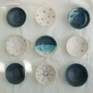 Op de afbeelding: Negen kleine, handgemaakte keramische kommen gerangschikt op een wit oppervlak. De kommen hebben verschillende ontwerpen, waaronder effen kleuren, gespikkelde patronen en abstracte blauw-witte ontwerpen. De kommen zijn in tinten blauw, wit en crème.