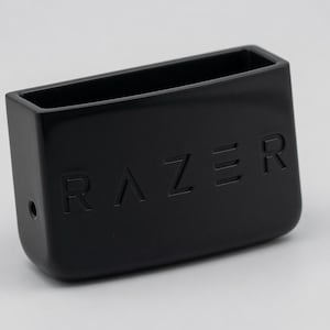 May include: A black rectangular container with rounded bottom corners and an open top. The front face features the embossed text RAZER in capital letters. A small circular hole is visible on the left side of the black plastic housing.