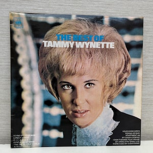 Peut inclure: Une pochette de disque vinyle vintage avec un portrait de Tammy Wynette, une chanteuse de musique country. La pochette est de couleur bleu clair avec une bordure blanche et porte le texte "The Best of Tammy Wynette".