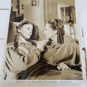 May include: Black and white publicity photograph for the film "Devotion" starring Ida Lupino and Paul Henreid. The photo shows the two actors in a close-up shot, looking at each other.