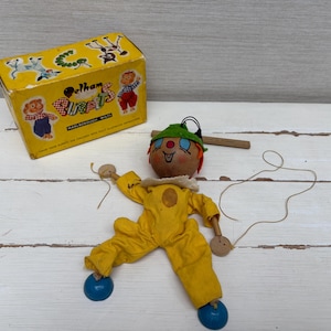 May include: A vintage Pelham Puppets marionette featuring a wooden head with a green hat, a yellow fabric jumpsuit, and blue circular feet. The puppet includes control strings and is accompanied by its original yellow cardboard box with illustrations.