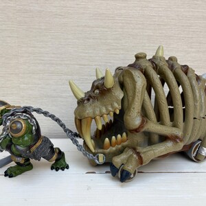 May include: A green and black fantasy figure pulls a bone-like cart with a large, toothy monster head. The cart has a ribcage design and wheels. The figure wears armor and a helmet, holding a sword.