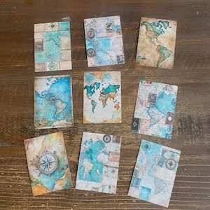 May include: Eight rectangular cards featuring vintage-style world map illustrations in shades of blue, tan, and brown. The designs include detailed cartographic elements, compass roses, and aged parchment textures, arranged in a grid on a dark wood surface.