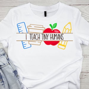 Könnte beinhalten: Weißes T-Shirt mit einem Grafikdesign aus einem roten Apfel, einem gelben Bleistift, einem blauen Lineal und einer braunen Kaffeetasse. Der Text "I teach tiny humans" ist auf einem schwarzen Banner gedruckt.