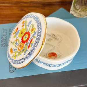 May include: A white porcelain trinket box with a floral design in shades of red, yellow, and blue. The lid is open and a silver chain with a red stone pendant is inside.