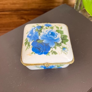 Puede incluir: Una caja de porcelana blanca con un borde dorado y un diseño floral azul con rosas.