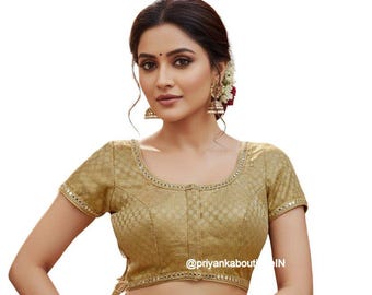 May include: A gold-coloured, short-sleeved blouse with a textured pattern and a rounded neckline. The neckline and sleeve edges are trimmed with small, reflective embellishments. The blouse is part of a traditional Indian outfit.
