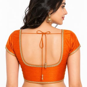 May include: Orange short-sleeved blouse with gold trim along the neckline and sleeves. The back features a tie closure and decorative gold beads. The blouse is made of a textured fabric and is likely part of a traditional garment.