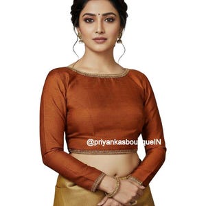 May include: A long-sleeved, rust-colored blouse with gold trim around the neckline and cuffs. The blouse is paired with a gold-colored skirt. The text "@priyankasboutiqueIN" is visible on the blouse.