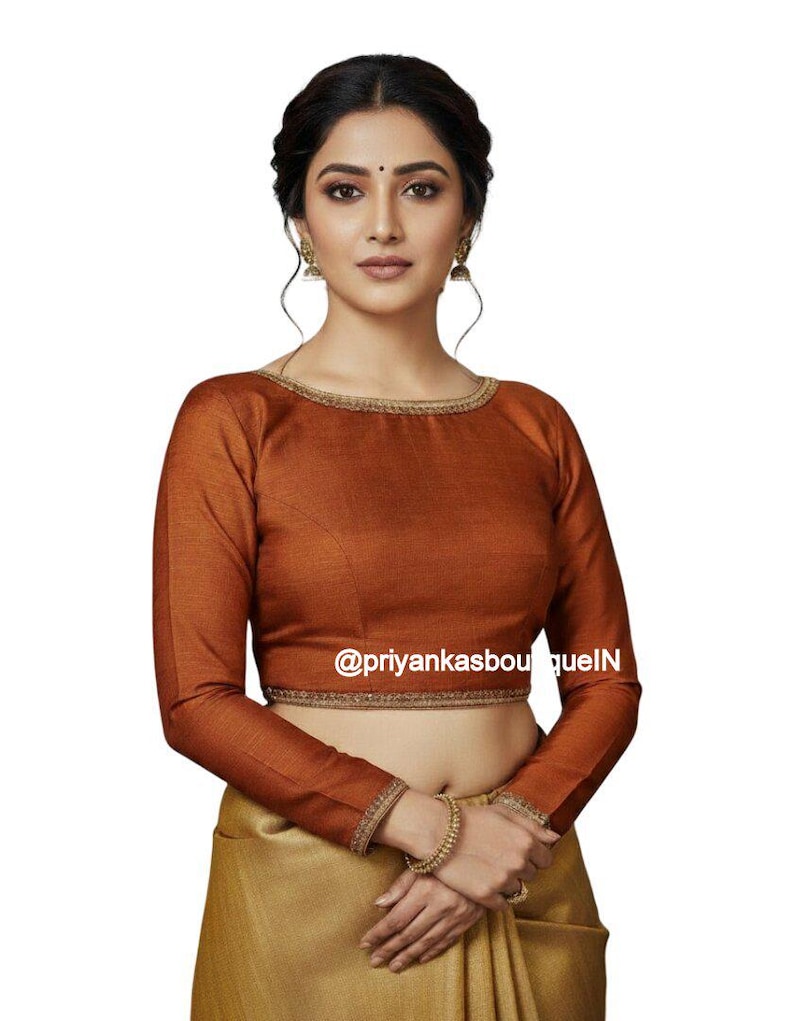 May include: A long-sleeved, rust-colored blouse with gold trim around the neckline and cuffs. The blouse is paired with a gold-colored skirt. The text "@priyankasboutiqueIN" is visible on the blouse.
