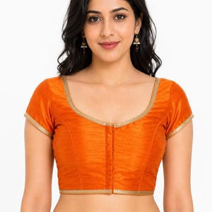 May include: An orange blouse with short sleeves and a gold trim. The blouse has a V-neckline and a vertical seam down the front. The fabric appears to be a smooth, possibly silk-like material. The blouse is part of a traditional Indian outfit.