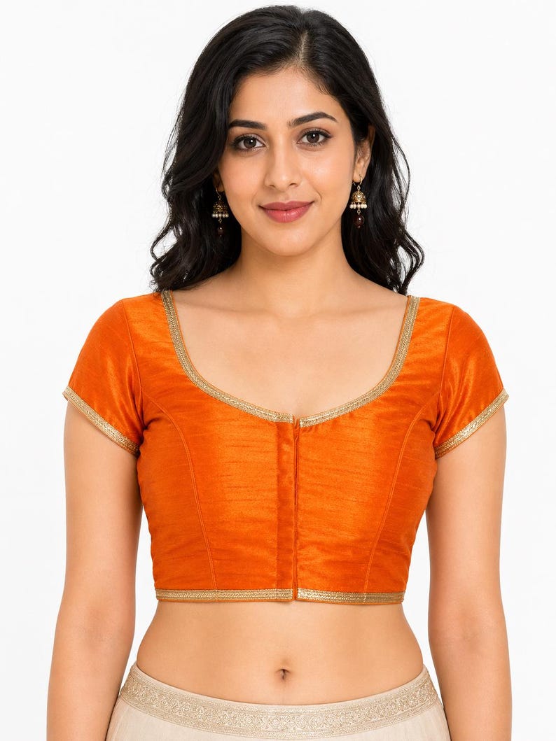May include: An orange blouse with short sleeves and a gold trim. The blouse has a V-neckline and a vertical seam down the front. The fabric appears to be a smooth, possibly silk-like material. The blouse is part of a traditional Indian outfit.