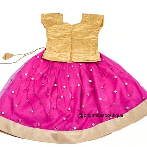 Op de afbeelding: Een roze en gouden meisjesoutfit met een gouden top en een roze rok met spiegelversieringen. De rok heeft een gouden zoom en een strik in de taille. De outfit is perfect voor een speciale gelegenheid.