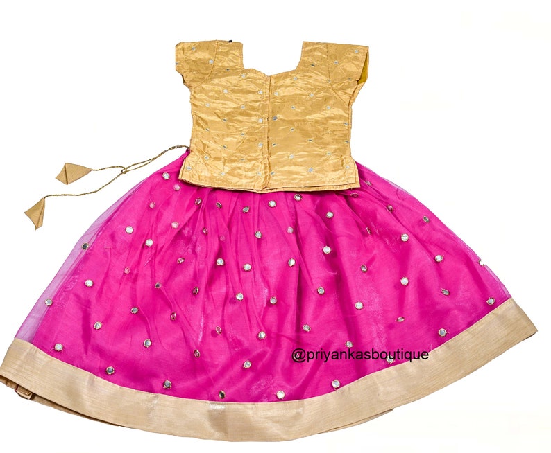 Op de afbeelding: Een roze en gouden meisjesoutfit met een gouden top en een roze rok met spiegelversieringen. De rok heeft een gouden zoom en een strik in de taille. De outfit is perfect voor een speciale gelegenheid.