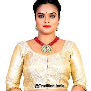 May include: A gold brocade blouse with a scoop neck and three-quarter sleeves. The blouse has a floral pattern and a button closure in the front. The blouse is worn by a person with a red bindi on their forehead and a red and green necklace.