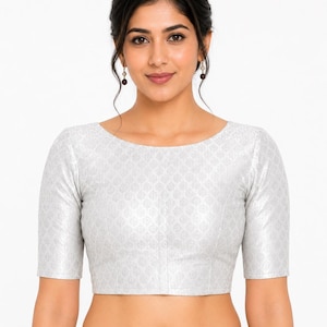 May include: A silver-colored, short-sleeved blouse with a subtle, repeating pattern. The blouse has a boat neckline and is cropped at the waist. The fabric appears to have a slight sheen. The model is wearing earrings.