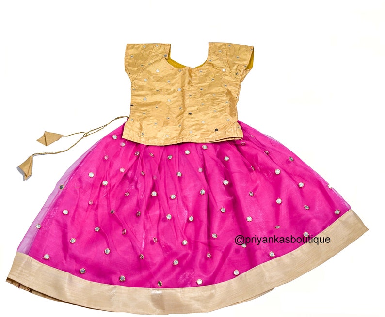 Op de afbeelding: Een roze en gouden meisjesoutfit met een kort, gouden topje en een roze rok met spiegelversieringen. De rok heeft een gouden zoom en een bijpassende gouden strik.