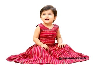 May include: A sleeveless red dress with a V-neck and a full skirt. The dress features a vertical striped pattern in shades of red and gold. The text "@priyankasboutiqueIN" is visible on the skirt. The dress is designed for a baby or toddler.