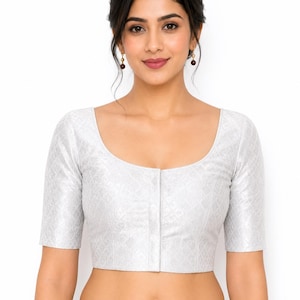Könnte beinhalten: Eine silberfarbene Bluse mit kurzen Ärmeln und einem dezenten geometrischen Muster. Die Bluse hat einen Rundhalsausschnitt und eine Knopfleiste vorne. Das Kleidungsstück ist Teil eines traditionellen indischen Outfits.
