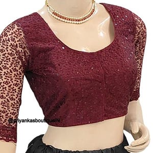 Op de afbeelding: Een bordeauxrode paillettenblouse met doorschijnende mouwen met bloemmotief. De top met ronde hals wordt op een mannequin gepresenteerd. De mouwen zijn driekwart. De blouse heeft een diepe bordeauxrode kleur.