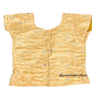 Op de afbeelding: Een gouden, mouwloze blouse met een ronde hals en korte mouwen. De blouse is versierd met kleine ronde spiegelversieringen. De tekst "@priyankasboutique" staat gedrukt in de rechterbenedenhoek van de blouse.