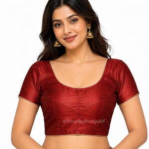 May include: A maroon-colored, short-sleeved blouse with a scoop neckline and gold trim. The blouse has a subtle sheen and is likely made of a smooth fabric. The text "@PriyankasBoutiqueIN" is visible on the lower part of the blouse.