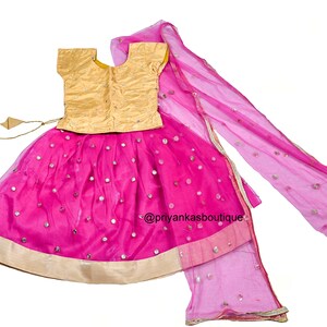 Op de afbeelding: Een roze en gouden meisjesoutfit met een rok, top en dupatta. De rok is versierd met kleine ronde spiegels en heeft een gouden rand. De top is goudkleurig met een ronde hals en korte mouwen. De dupatta is een doorschijnende roze stof met een gouden rand.