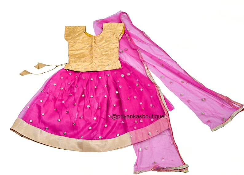 Op de afbeelding: Een roze en gouden meisjesoutfit met een rok, top en dupatta. De rok is versierd met kleine ronde spiegels en heeft een gouden rand. De top is goudkleurig met een ronde hals en korte mouwen. De dupatta is een doorschijnende roze stof met een gouden rand.