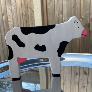 May include: A wooden cow figurine painted white with black spots and pink accents. The cow is standing on a silver tray.