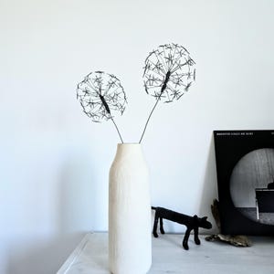 Könnte beinhalten: Zwei schwarze Drahtskulpturen in Form von Löwenzahn-Samenköpfen mit dünnen Stielen, platziert in einer hohen, zylindrischen, cremeweißen Keramikvase mit einem dezenten vertikalen Strukturmuster auf der Oberfläche, stehend vor einer schlichten Wand.