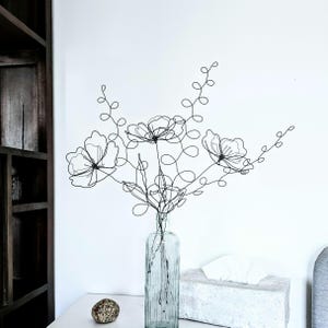 Può includere: Una scultura di fiori in filo di ferro in una bottiglia di vetro trasparente. I fiori in filo di ferro nero hanno un design delicato e aperto, con steli e petali ad anello. La bottiglia è su una superficie bianca.