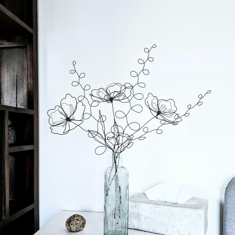May include: A black wire sculpture shaped like a bouquet of three flowers with curly stems and leaves, placed inside a clear, vertically ribbed glass bottle. The wire art features delicate, continuous lines forming petals and stems.
