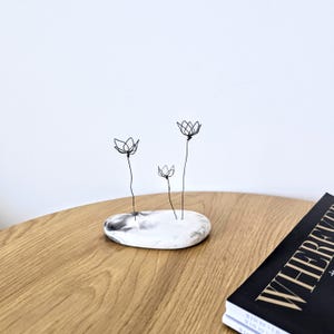 Pode incluir: Uma escultura decorativa com três flores de arame com caules e pétalas pretas. As flores estão colocadas numa base oval branca e cinzenta. A escultura é exibida numa mesa de madeira.