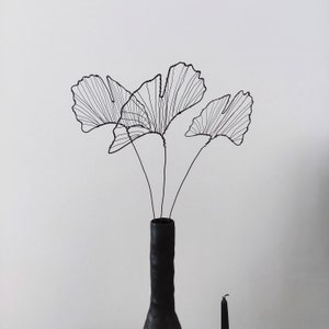 Op de afbeelding: Een zwarte vaas met een getextureerd oppervlak houdt drie draadbeelden van ginkgo bladeren vast.