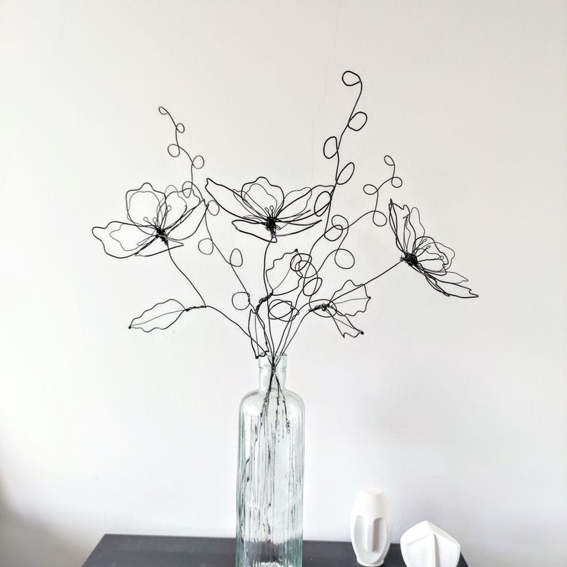 May include: A sculpture of flowers crafted from thin black wire, featuring three distinct blossoms with delicate petals, winding stems, and curled tendrils. The wire floral arrangement is displayed inside a clear, vertically ribbed glass bottle.