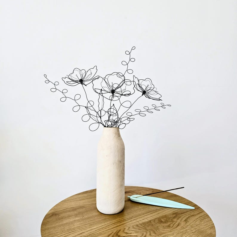 May include: A black wire sculpture shaped like a bouquet of flowers with swirling stems and petals, placed inside a tall, cylindrical, light-colored wooden vase. A pale blue, feather-shaped incense holder rests on the surface nearby.