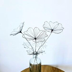 Könnte beinhalten: Ein dekoratives Arrangement aus drei schwarzen Drahtblumen mit zarten, dünnen Blütenblättern und Stielen, ergänzt durch filigranes Drahtlaub. Die Drahtskulptur wird in einer klaren Glasflasche präsentiert und zeigt die feine Handwerkskunst.
