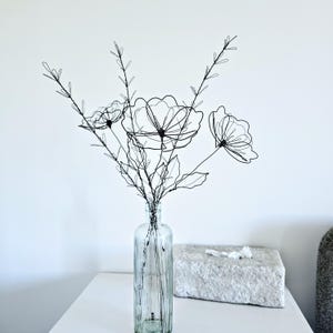 May include: A decorative arrangement of black wire flowers and stems placed inside a clear glass bottle. The sculpture features three distinct flower heads with delicate wire petals and thin, branching stems with small wire leaves.