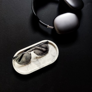 Könnte beinhalten: Ein ovales Tablett aus weißem Marmor mit einer schwarzen Sonnenbrille darauf. Die Sonnenbrille hat dunkelgraue Gläser. Das Tablett befindet sich auf einer schwarzen Oberfläche.