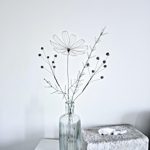 Könnte beinhalten: Eine schwarze Drahtskulptur einer einzelnen großen Blume mit mehreren Blütenblättern und mehreren dünnen Stielen mit kleinen runden Knospen und zarten blattartigen Strukturen, angeordnet in einer klaren, vertikal gerippten Glasflasche.