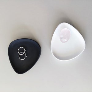 May include: Two small, triangular ceramic dishes. One is black and holds two silver rings. The other is white and holds a pink crystal.